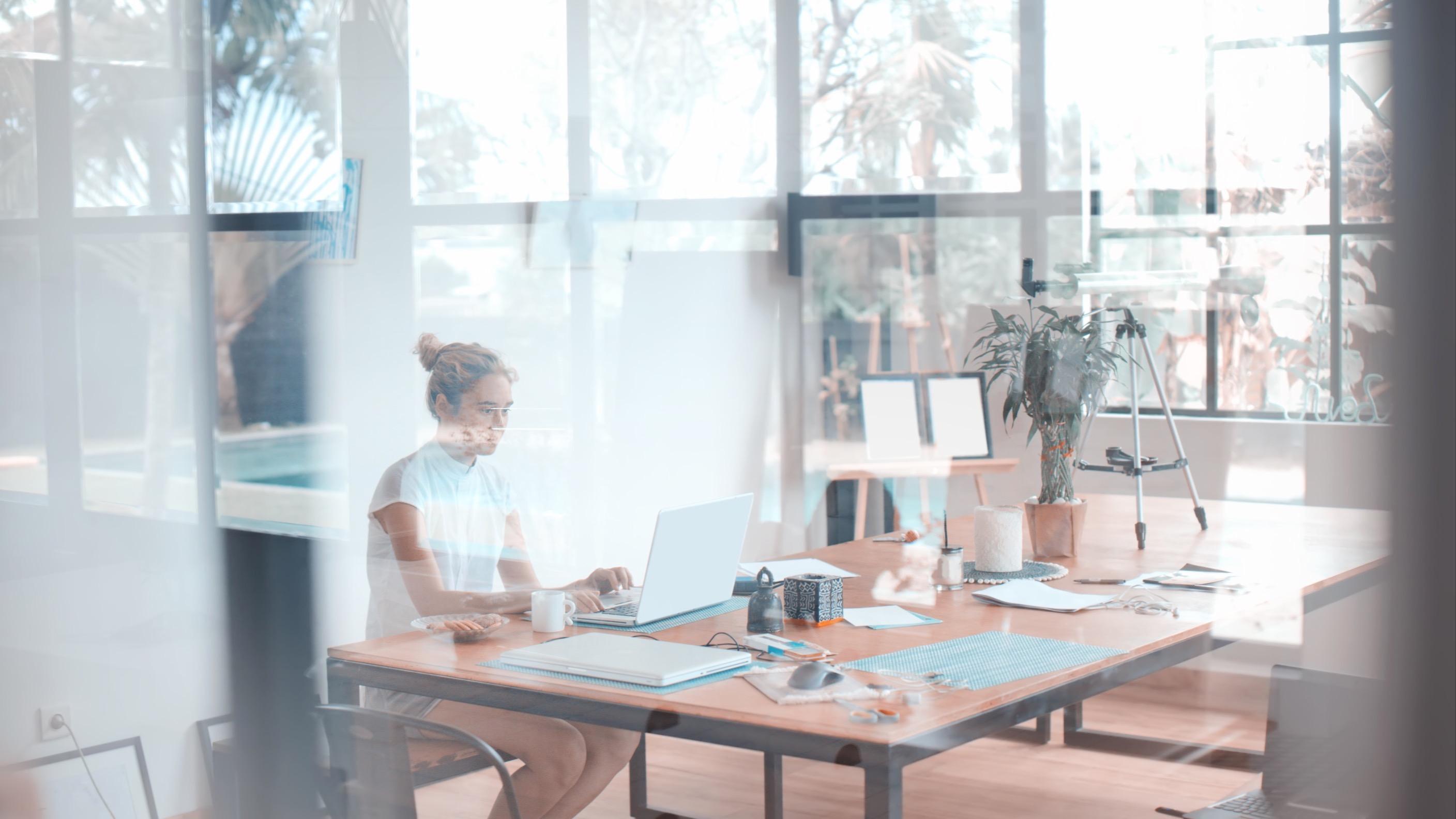 Woman sitting using her laptop at a table behind glass wall