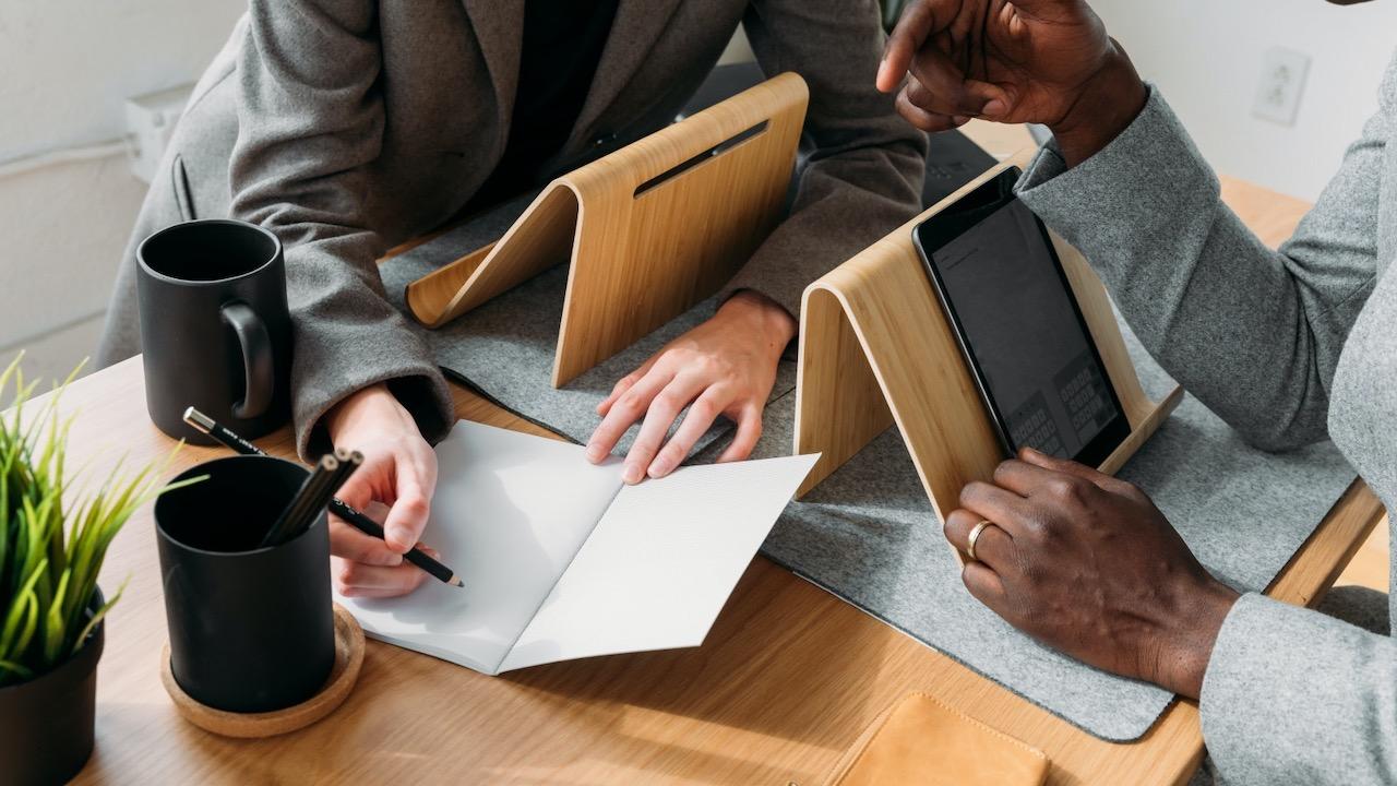 Two people talking and looking at a notebook at a desk with tablets