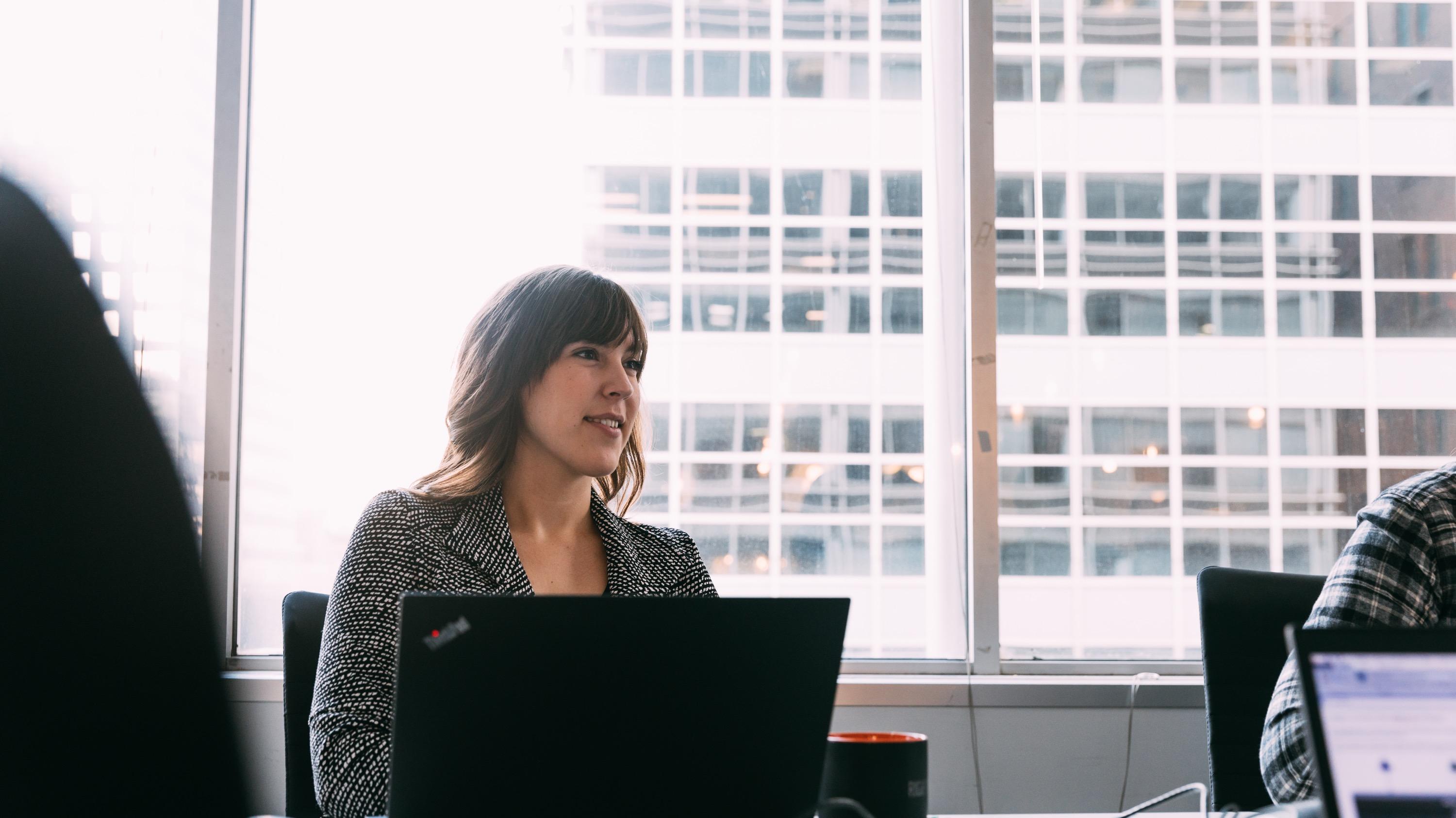 Woman sitting behind a laptop at a conference table with a window behind her