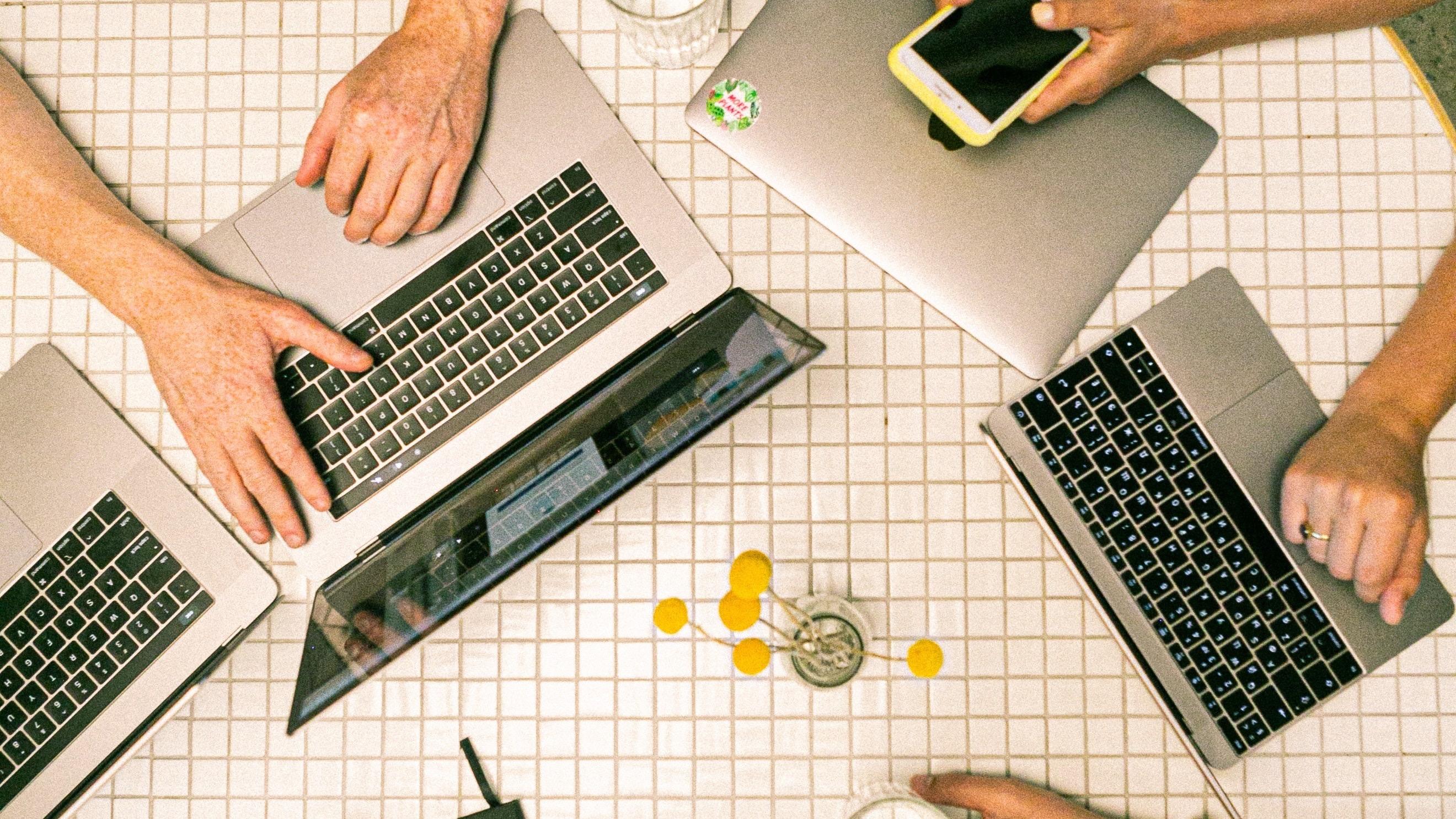 Birds eye view of people sitting at a round table with phones, laptops, tablets, and notebooks